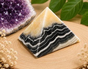 May include: A layered stone pyramid with black, white, and yellow bands sits on a wooden surface. A purple amethyst geode is in the background. The pyramid is a decorative item.