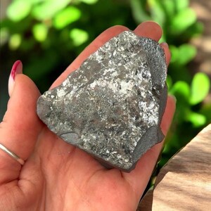 K&ouml;nnte beinhalten: Ein dunkelgraues, unregelm&auml;&szlig;ig geformtes Mineralexemplar mit metallischem Glanz. Die Oberfl&auml;che weist Bereiche aus silberfarbenem, reflektierendem Material auf. Der Stein wird in einer Hand gehalten, mit einem verschwommenen gr&uuml;nen Hintergrund.