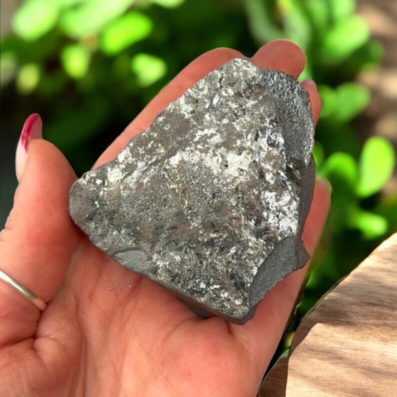 K&ouml;nnte beinhalten: Ein dunkelgraues, unregelm&auml;&szlig;ig geformtes Mineralexemplar mit metallischem Glanz. Die Oberfl&auml;che weist Bereiche aus silberfarbenem, reflektierendem Material auf. Der Stein wird in einer Hand gehalten, mit einem verschwommenen gr&uuml;nen Hintergrund.
