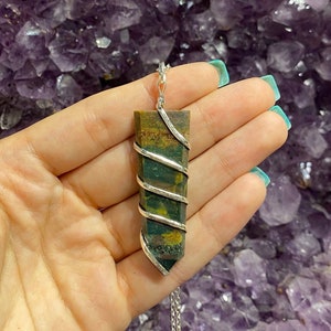 May include: A rectangular pendant with a silver wire wrap and chain. The pendant features green, brown, and yellow tones. The background is a blurred purple colour.