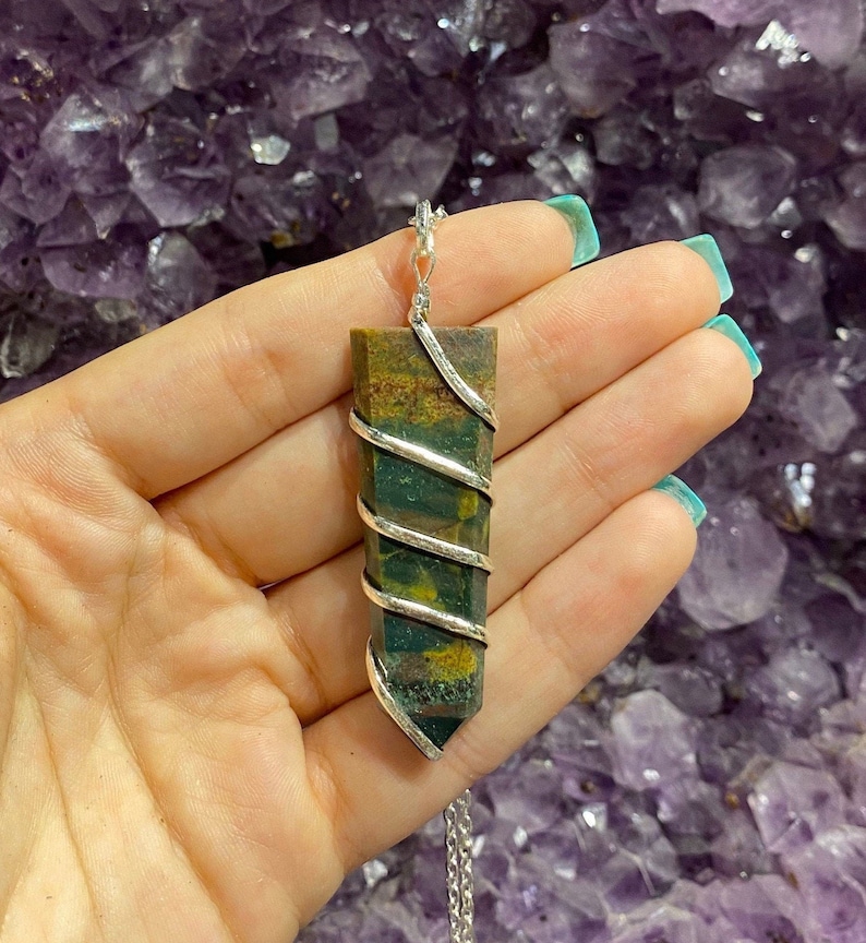 May include: A rectangular pendant with a silver wire wrap and chain. The pendant features green, brown, and yellow tones. The background is a blurred purple colour.