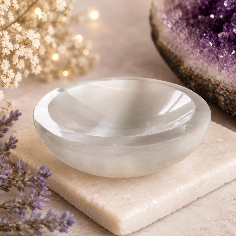 May include: A small, white selenite bowl with a smooth, polished surface. The bowl is oval-shaped and sits on a square, light-coloured stone. The background includes a purple amethyst geode and delicate white flowers.