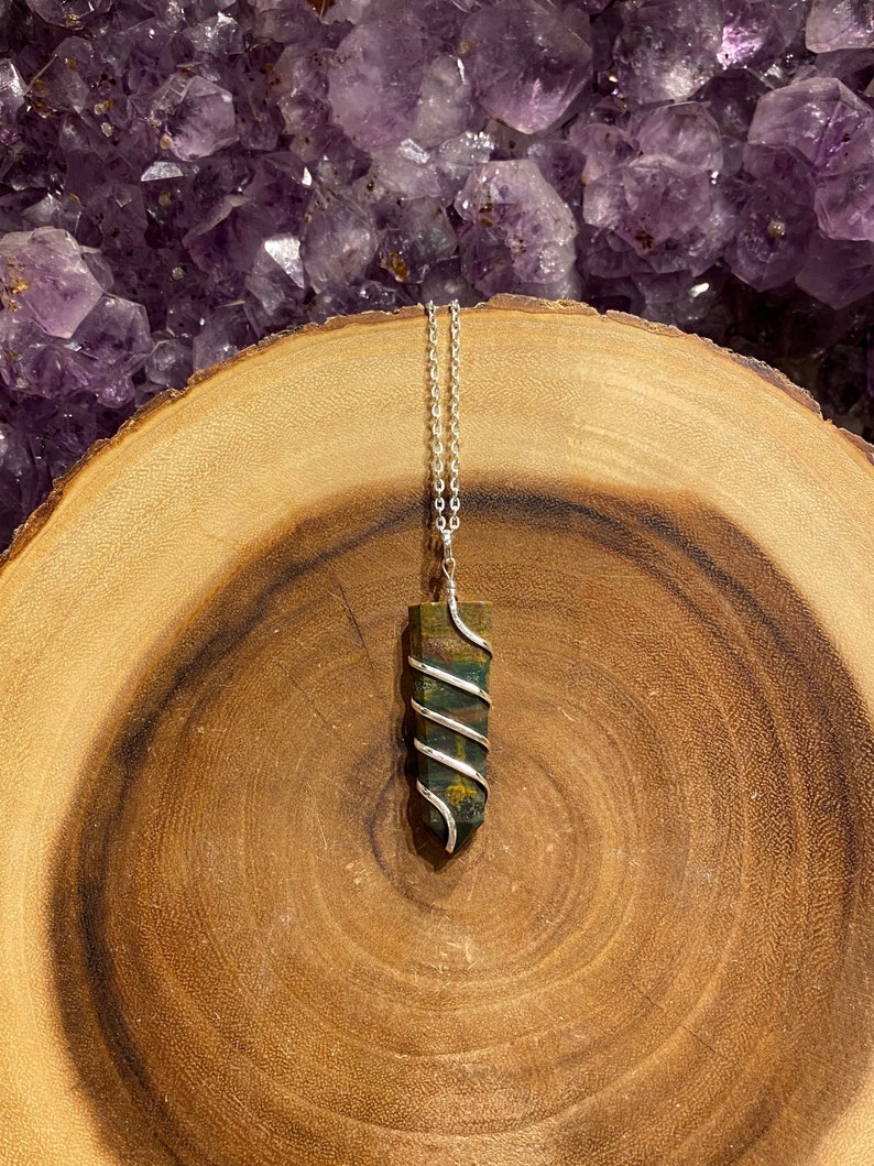 May include: A silver pendant necklace with a rectangular, multi-coloured stone wrapped in silver wire. The stone shows shades of green, brown, and red. The necklace is displayed on a wooden surface with a purple crystal background.