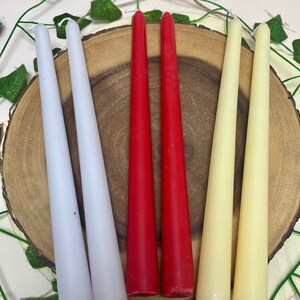May include: A set of six taper candles in three colors: white, red, and cream. The candles are arranged on a wooden surface.