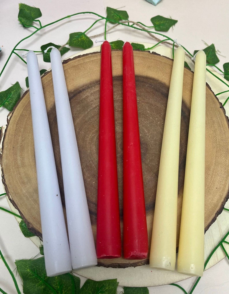 May include: A set of six taper candles in three colors: white, red, and cream. The candles are arranged on a wooden surface.