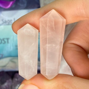 May include: Two rose quartz crystal points, one larger than the other, are held in a hand. The crystals are translucent and have a pale pink color.