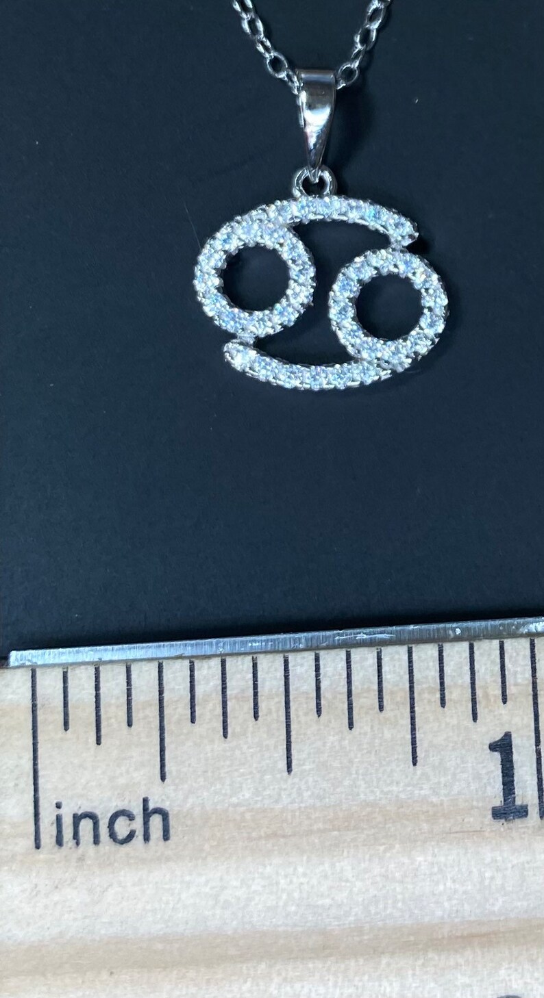 May include: A silver pendant necklace with a clear crystal Cancer zodiac sign charm. The pendant is about 1 inch in size.