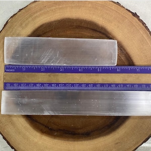 May include: Two rectangular, translucent selenite crystal wands are displayed on a wooden surface. A purple ruler with centimetre measurements is placed next to the crystals. The longer wand is approximately 25 cm long.