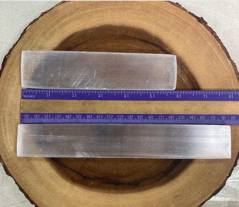 May include: Two rectangular, translucent selenite crystal wands are displayed on a wooden surface. A purple ruler with centimetre measurements is placed next to the crystals. The longer wand is approximately 25 cm long.