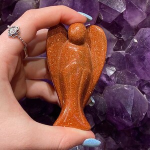 May include: A small, orange-brown, polished, angel-shaped, gemstone sculpture. The sculpture is made of sunstone and has a sparkly, glittery finish.