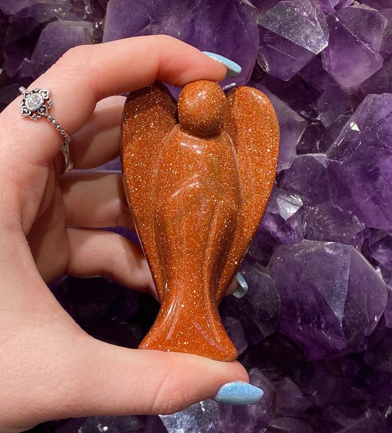 May include: A small, orange-brown, polished, angel-shaped, gemstone sculpture. The sculpture is made of sunstone and has a sparkly, glittery finish.