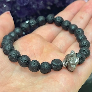 May include: A black lava rock beaded bracelet with a silver metal charm. The charm is a rough, irregular shape and appears to be made of a meteorite.