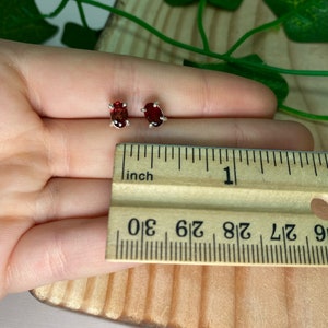May include: A pair of silver stud earrings with oval-shaped red gemstones. The earrings are shown next to a ruler, with the gemstones measuring approximately 1/4 inch in length.