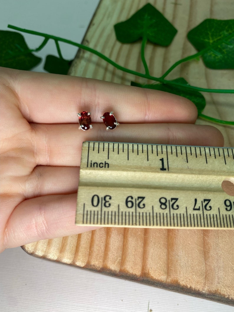 May include: A pair of silver stud earrings with oval-shaped red gemstones. The earrings are shown next to a ruler, with the gemstones measuring approximately 1/4 inch in length.