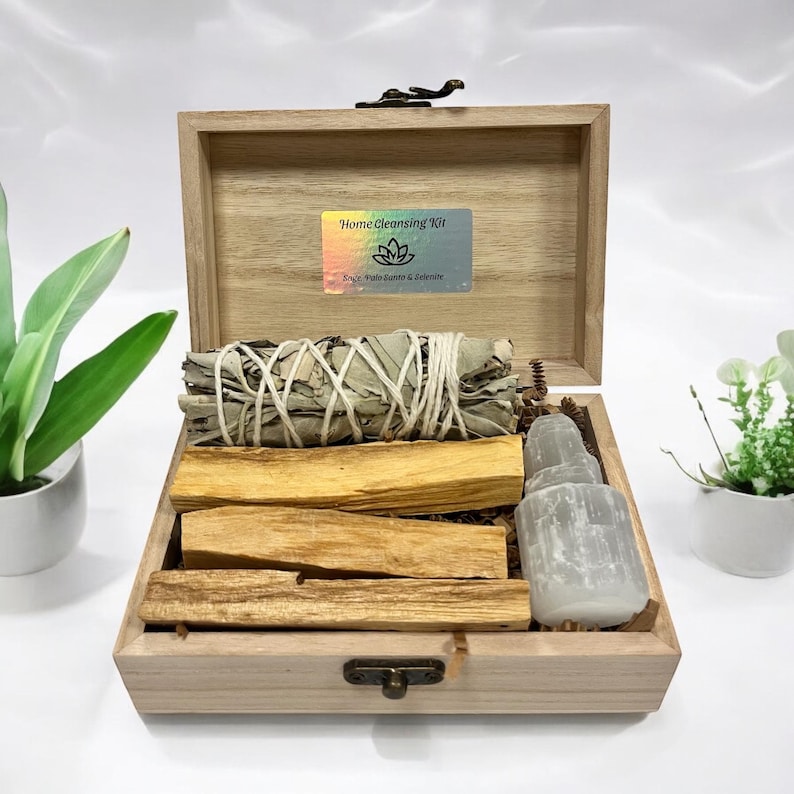 May include: A wooden box with a lid open, revealing a home cleansing kit. The kit includes a bundle of white sage, palo santo sticks, and a clear crystal. The box has a label that reads "Home Cleansing Kit" with a lotus flower symbol and the text "Sage, Palo Santo & Selenite".