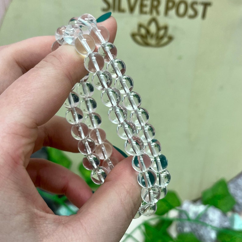 May include: Clear quartz crystal bead bracelets. The bracelets are made of round, transparent quartz beads strung together. The beads are held in a person's hand, with the background showing the text "SILVER POST".