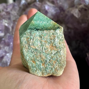 May include: A green and brown crystal point with a rough, textured surface. The crystal is a natural, raw, and unpolished stone.