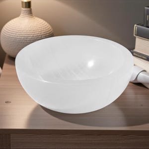 May include: A white, translucent, round bowl with a smooth surface. The bowl is on a wooden surface. A decorative lamp and books are in the background.