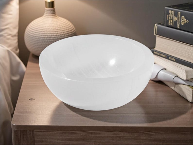 May include: A white, translucent, round bowl with a smooth surface. The bowl is on a wooden surface. A decorative lamp and books are in the background.