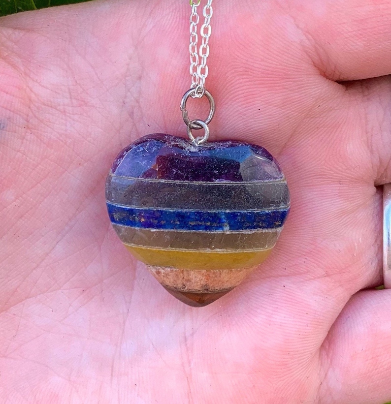 K&ouml;nnte beinhalten: Eine silberne Kettenkette mit einem herzf&ouml;rmigen Anh&auml;nger aus geschichteten Edelsteinen. Der Anh&auml;nger besteht aus sieben Schichten verschiedener farbiger Steine, darunter Amethyst, klarer Quarz, Lapislazuli, Tigerauge, Citrin, Karneol und roter Jaspis.