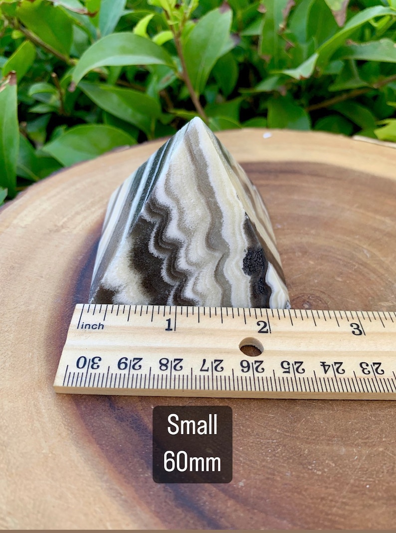 May include: A small, pyramid-shaped stone with a layered pattern of black, tan, and cream colours. The stone is approximately 5 cm in height. The text "Small 60mm" is visible.