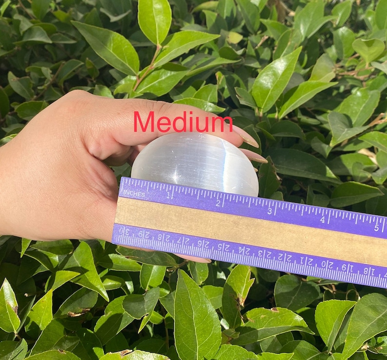 K&ouml;nnte beinhalten: Eine klare Selenitkugel mit einem Durchmesser von etwa 5 cm wird in einer Hand gehalten. Die Kugel liegt auf einem Lineal, &uuml;ber dem der Text "Medium" zu sehen ist.