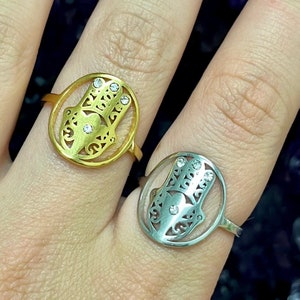 Puede incluir: Dos anillos, uno dorado y otro plateado, cada uno con un diseño de mano de Hamsa dentro de un marco circular. Las manos de Hamsa tienen intrincados patrones calados y pequeñas gemas transparentes. Los anillos se muestran en un dedo.