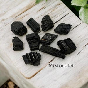 May include: A lot of ten black tourmaline stones, also known as schorl, on a white wooden surface.  The stones are rough and have a natural, unpolished finish.  10 stone lot.