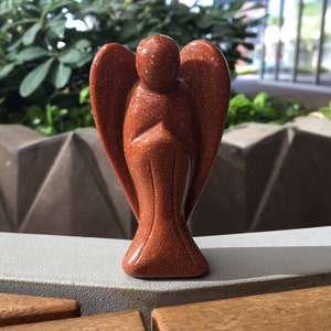 May include: A small, red-brown sandstone angel figurine. The angel has wings and is standing with its hands clasped in front of it.