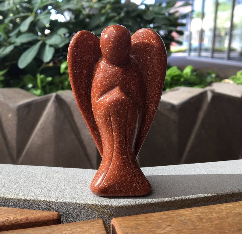 May include: A small, red-brown sandstone angel figurine. The angel has wings and is standing with its hands clasped in front of it.