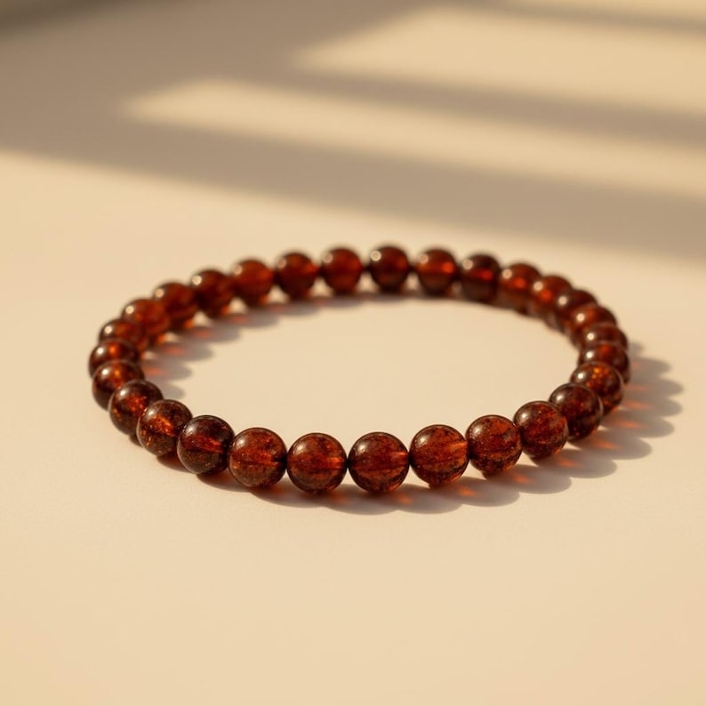 May include: A beaded bracelet featuring small, round, reddish-brown beads. The bracelet is circular and rests on a light-colored surface. The beads appear to be evenly sized and spaced, creating a smooth, continuous loop.