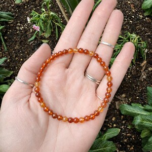 May include: A beaded bracelet featuring small, round, orange and yellow gemstones. The bracelet is displayed on a hand with several silver rings. The background shows greenery and soil.