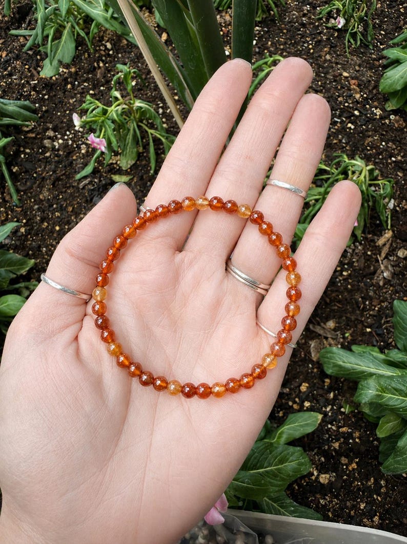 May include: A beaded bracelet featuring small, round, orange and yellow gemstones. The bracelet is displayed on a hand with several silver rings. The background shows greenery and soil.
