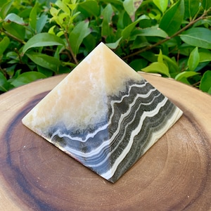 May include: A decorative pyramid ornament with a layered design. The top half is a creamy beige colour, while the bottom half features bands of black, grey, and white. The pyramid is displayed on a wooden surface with green foliage in the background.