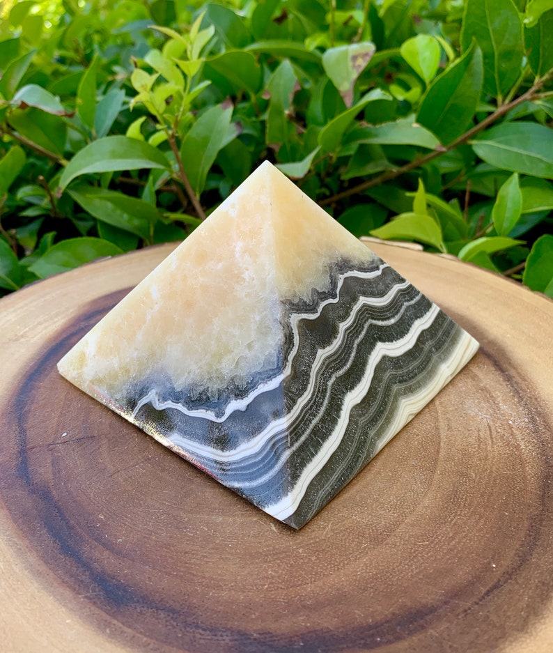 May include: A decorative pyramid ornament with a layered design. The top half is a creamy beige colour, while the bottom half features bands of black, grey, and white. The pyramid is displayed on a wooden surface with green foliage in the background.
