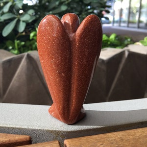 May include: A red sandstone angel figurine with a sparkly finish. The angel is standing on a light grey surface.