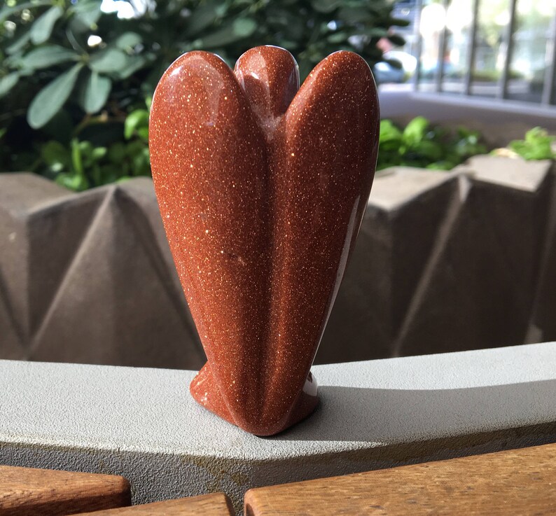 May include: A red sandstone angel figurine with a sparkly finish. The angel is standing on a light grey surface.
