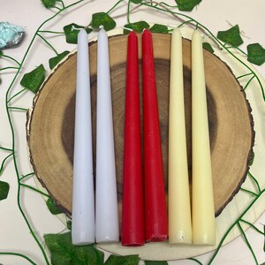 May include: Six taper candles in three colors: white, red, and cream. The candles are arranged in a row on a wooden surface.