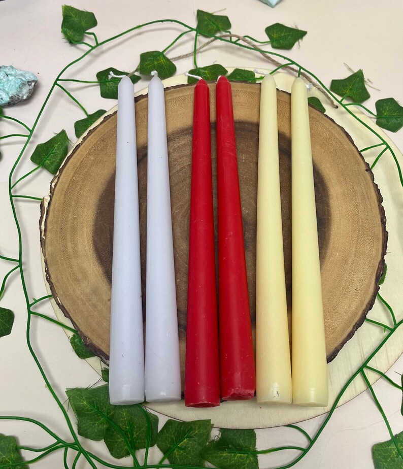 May include: Six taper candles in three colors: white, red, and cream. The candles are arranged in a row on a wooden surface.