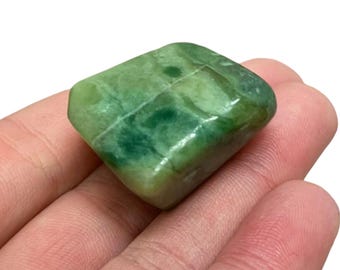 May include: A polished, green jade stone with a smooth, rounded shape. The stone has varying shades of green, with darker green veining throughout. The jade is held in a hand against a white background.
