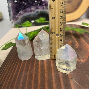 K&ouml;nnte beinhalten: Drei regenbogenfarbene, klare Quarzkristallspitzen, jede etwa 5 cm hoch, sind auf einer Holzoberfl&auml;che angeordnet. Ein Lineal dient zur Veranschaulichung der Gr&ouml;&szlig;e der Kristalle.