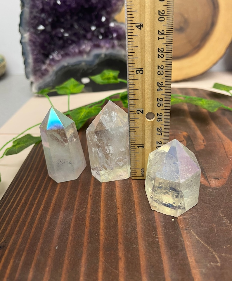 K&ouml;nnte beinhalten: Drei regenbogenfarbene, klare Quarzkristallspitzen, jede etwa 5 cm hoch, sind auf einer Holzoberfl&auml;che angeordnet. Ein Lineal dient zur Veranschaulichung der Gr&ouml;&szlig;e der Kristalle.