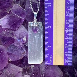 May include: A silver chain necklace with a white selenite crystal pendant and a small amethyst crystal charm. The pendant is approximately 1.75 inches long. The necklace is displayed on a bed of amethyst crystals.