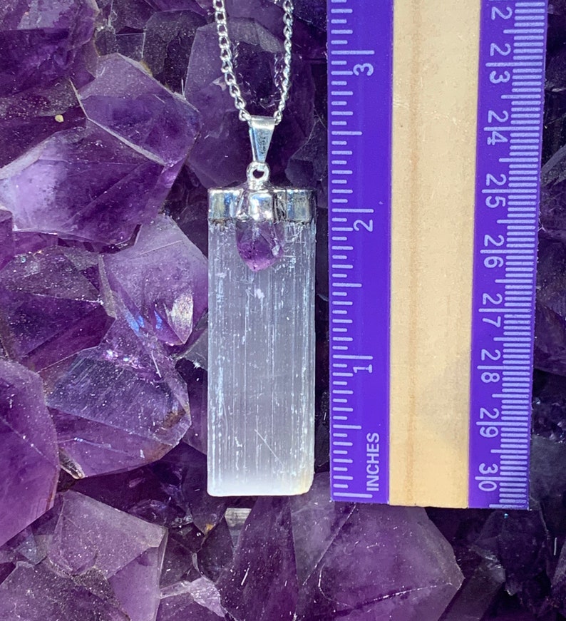May include: A silver chain necklace with a white selenite crystal pendant and a small amethyst crystal charm. The pendant is approximately 1.75 inches long. The necklace is displayed on a bed of amethyst crystals.