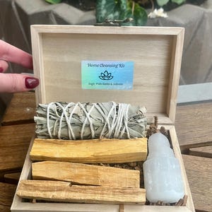 May include: A wooden box with a hinged lid containing a home cleansing kit. The kit includes sage, palo santo, and selenite. The box is labeled "Home Cleansing Kit" with the text "Sage, Palo Santo & Selenite".