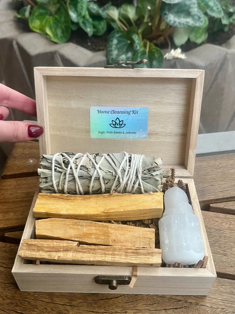 May include: A wooden box with a hinged lid containing a home cleansing kit. The kit includes sage, palo santo, and selenite. The box is labeled "Home Cleansing Kit" with the text "Sage, Palo Santo & Selenite".