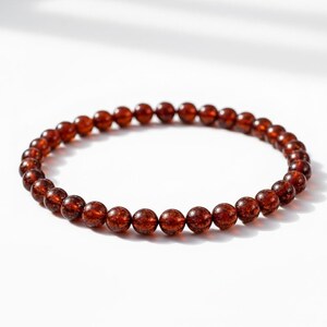May include: A beaded bracelet featuring small, round, reddish-brown beads. The beads are strung together to form a circular shape, resting on a white surface. The bracelet is a simple, elegant piece of jewelry.
