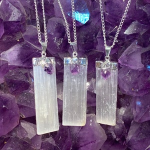 May include: Three silver necklaces with selenite crystal pendants and amethyst accents. The pendants are rectangular and have a white, striated appearance. The necklaces are displayed against a background of amethyst crystals.
