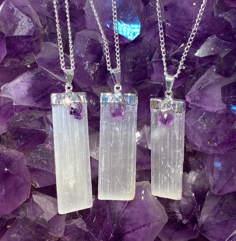 May include: Three silver necklaces with selenite crystal pendants and amethyst accents. The pendants are rectangular and have a white, striated appearance. The necklaces are displayed against a background of amethyst crystals.