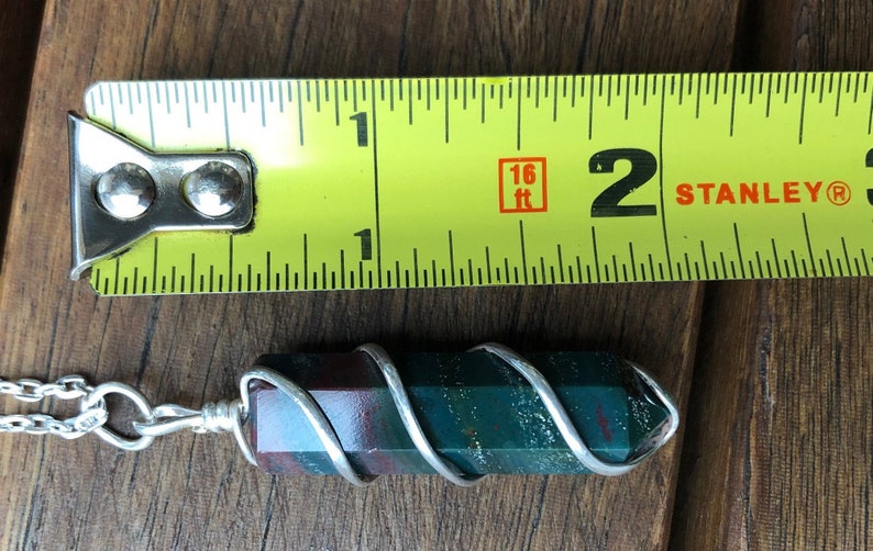 May include: A green, red, and brown stone pendant wrapped in silver wire on a silver chain. The pendant is approximately 2 inches long.